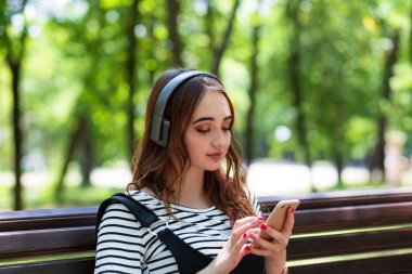 Genç, güzel, kızıl saçlı bir kız bir bankta otururken kulaklıkla müzik dinliyor.