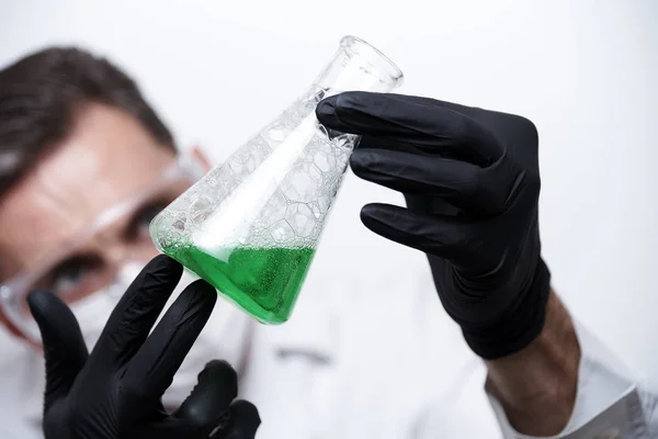 A flask with a green liquid and bubbles in the hands of a young man. The scientist in uniform in protective glasses, a mask and black gloves conducts an experiment.