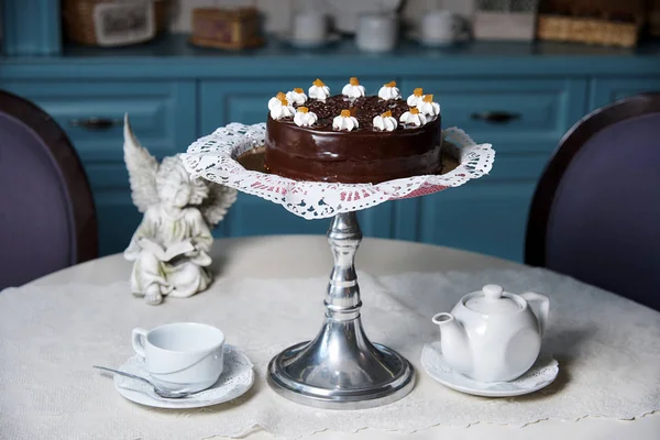 A chocolate cake on a stand stands on a decorated table with lace with a cup, a teapot and an angel statue.