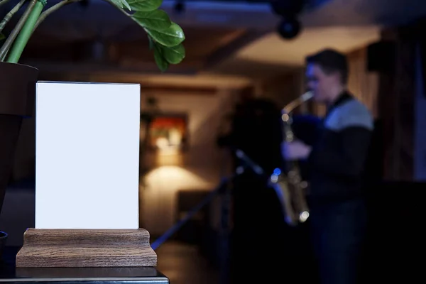 Mockup label of an empty frame menu in a restaurant bar, stand for booklets with white sheets of paper against a blurred background of a saxophonist playing in the twilight.