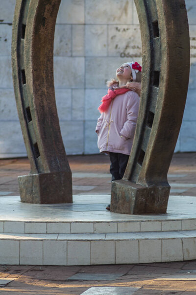 Маленькая девочка на весенней прогулке у памятника в виде подковы, приносящей счастье
