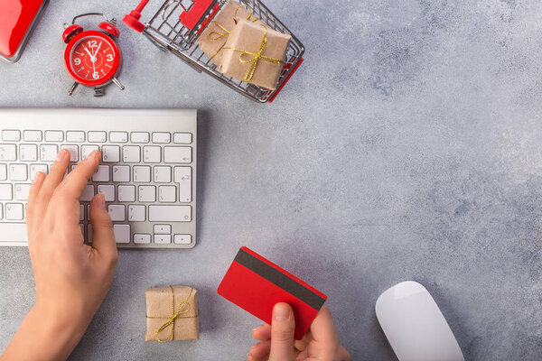 Woman hands ordering gifts online, paying by credit card. Concept online shopping buying presents. Red credit card, keyborad and christmas presents on grey table flat lay, copy space.