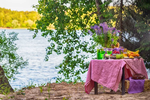 Piquenique perto da água à mesa com flores em frutas e legumes da floresta — Fotografia de Stock
