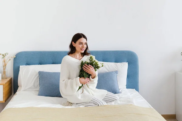 Una chica en un suéter blanco se sienta en una cama con un ramo de rosas blancas en la mano — Foto de Stock