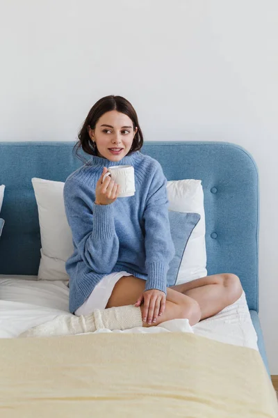 Chica en un suéter azul en el interior de estilo hygge con una taza de té caliente su mano se sienta en la cama — Foto de Stock