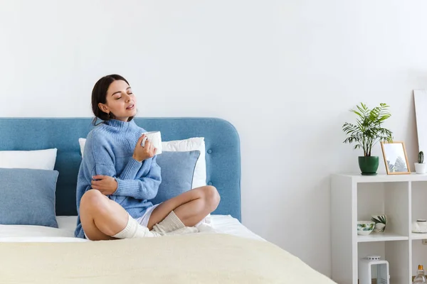 Chica en un suéter azul en el interior estilo Hygge con una taza en la mano se sienta en la cama — Foto de Stock