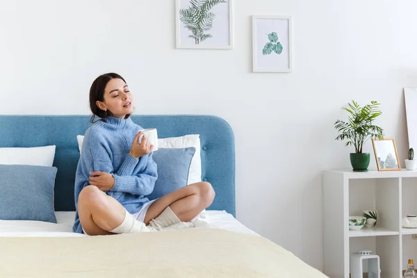 Chica en un suéter azul en el interior estilo Hygge con una taza en la mano se sienta en la cama — Foto de Stock