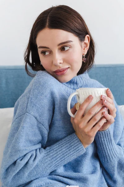 Piękna twarz dziewczyny, Dziewczyna w niebieskim swetrze siedzi na kanapie z filiżanką gorącej herbaty w dłoniach — Zdjęcie stockowe