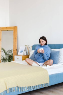 Elinde bir fincan ile iç Hygge tarzı mavi bir kazak Kız yatakta oturur
