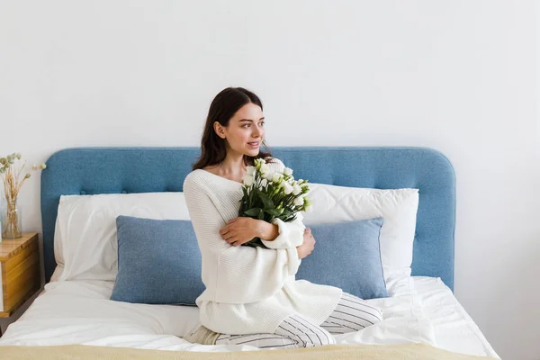Una chica en un suéter blanco se sienta en una cama con un ramo de rosas blancas en la mano — Foto de Stock