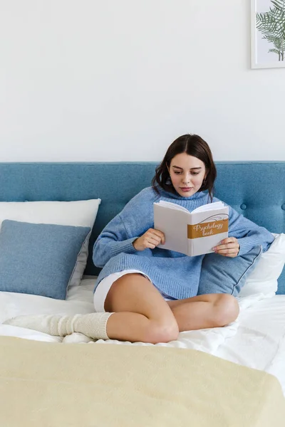 Chica en un suéter azul leyendo un libro sobre psicología sentado en la cama en un interior acogedor — Foto de Stock