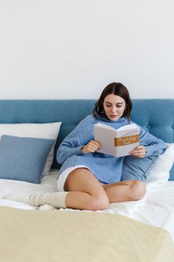 Mavi kazak giyen kız, rahat bir iç mekanda yatakta oturup psikoloji üzerine bir kitap okuyor.