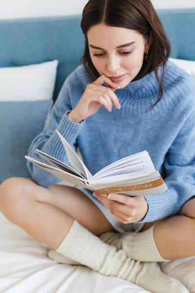 Gadis dengan sweater biru membaca buku tentang psikologi duduk di tempat tidur di interior nyaman — Stok Foto