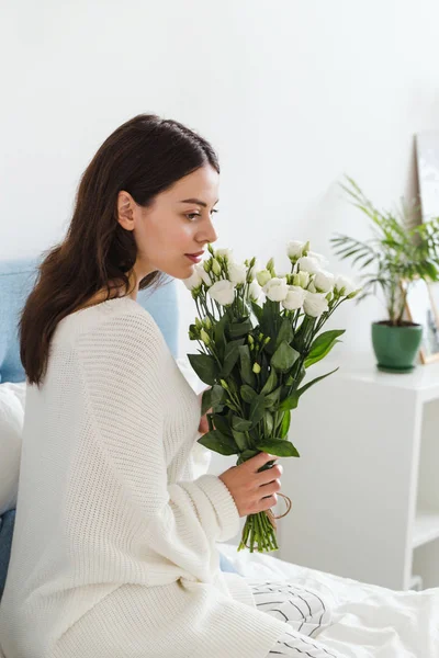 Una chica en un suéter blanco se sienta en una cama con un ramo de rosas blancas en la mano — Foto de Stock