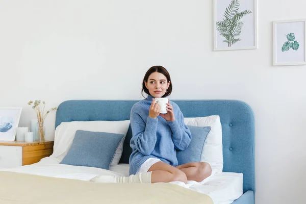 Chica en un suéter azul en el interior estilo Hygge con una taza en la mano se sienta en la cama — Foto de Stock