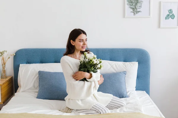 Una chica en un suéter blanco se sienta en una cama con un ramo de rosas blancas en la mano — Foto de Stock