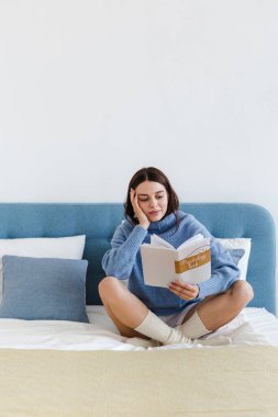 Mavi kazak giyen kız, rahat bir iç mekanda yatakta oturup psikoloji üzerine bir kitap okuyor.