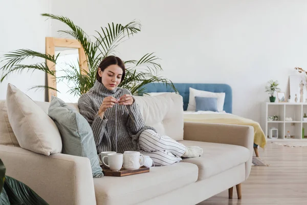 Chica en un suéter gris cálido teje sentado en un sofá en un acogedor interior hygge — Foto de Stock
