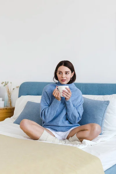 Chica en un suéter azul en el interior estilo Hygge con una taza en la mano se sienta en la cama — Foto de Stock