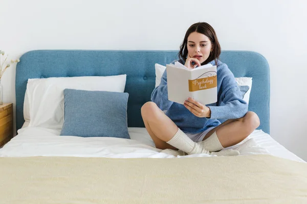 Girl in a blue sweater reading a book on psychology sitting on the bed in a cozy interior
