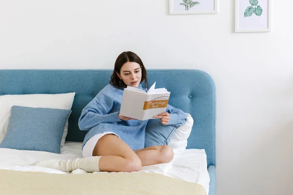 Chica en un suéter azul leyendo un libro sobre psicología sentado en la cama en un interior acogedor — Foto de Stock