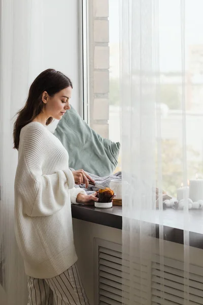 Una chica en un suéter blanco se para cerca de la ventana y corta una magdalena — Foto de Stock