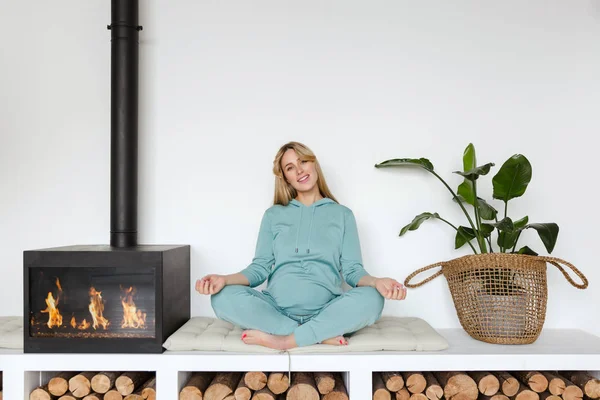Chica embarazada en ropa deportiva verde se sienta en posición de loto haciendo yoga sonriendo en el interior acogedor — Foto de Stock