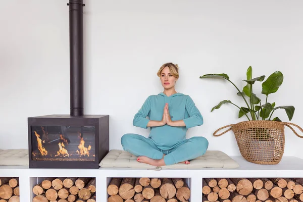 Chica embarazada en ropa deportiva azul se sienta en posición de loto haciendo yoga en interior acogedor — Foto de Stock