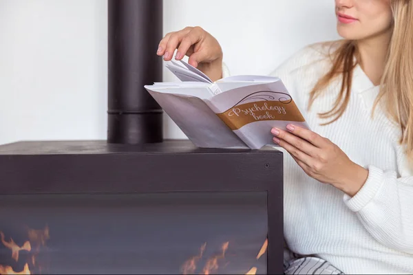 Rubia se sienta en un acogedor interior escandinavo junto a la chimenea y lee un libro sobre psicología — Foto de Stock