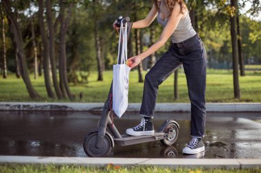 Bir esmer kız parkta elektrikli scooter yanında duruyor ve bir çanta alır