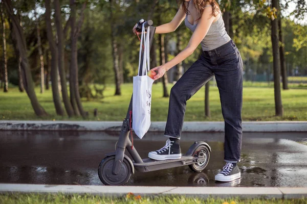 Брюнетка стоїть поруч з електричним скутером в парку і бере з сумки — стокове фото