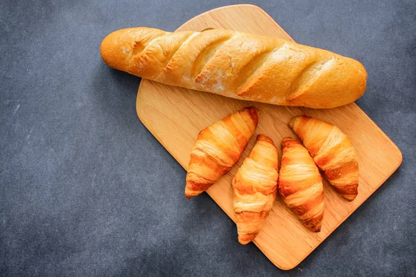 French loaf and four croissants on a wooden cutting board on dark craft paper.