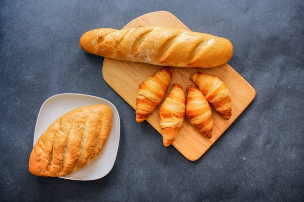 French loaf and four croissants on a wooden cutting board on dark craft paper.