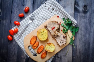 Kesme tahtası üzerinde pişmiş et iki dilim, iki dilim patates ve havuç dilimleri, keten, kareli bir peçete pişmiş bir kapta çok renkli bezelye biber ve grafit tezgahı çerçevede bir tabakta kesilmiş bir nar vardır