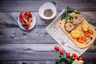 Kesme tahtası üzerinde pişmiş et iki dilim, iki dilim patates ve havuç dilimleri, keten, kareli bir peçete pişmiş bir kapta çok renkli bezelye biber ve grafit tezgahı çerçevede bir tabakta kesilmiş bir nar vardır