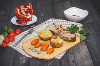 Kesme tahtası üzerinde pişmiş et iki dilim, iki dilim patates ve havuç dilimleri, keten, kareli bir peçete pişmiş bir kapta çok renkli bezelye biber ve grafit masa üstü karşı bir tabakta kesilmiş bir nar vardır