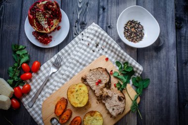 Kesme tahtası üzerinde pişmiş et iki dilim, iki dilim patates ve havuç dilimleri, keten, kareli bir peçete pişmiş bir kapta çok renkli bezelye biber ve grafit masa üstü karşı bir tabakta kesilmiş bir nar vardır