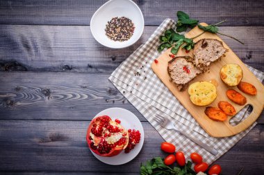 Kesme tahtası üzerinde pişmiş et iki dilim, iki dilim patates ve havuç dilimleri, keten, kareli bir peçete pişmiş bir kapta çok renkli bezelye biber ve grafit masa üstü karşı bir tabakta kesilmiş bir nar vardır