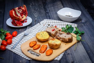 Kesme tahtası üzerinde pişmiş et iki dilim, iki dilim patates ve havuç dilimleri, keten, kareli bir peçete pişmiş bir kapta çok renkli bezelye biber ve grafit masa üstü karşı bir tabakta kesilmiş bir nar vardır