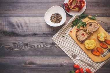 Kesme tahtası üzerinde pişmiş et iki dilim, iki dilim patates ve havuç dilimleri, keten, kareli bir peçete pişmiş bir kapta çok renkli bezelye biber ve grafit masa üstü karşı bir tabakta kesilmiş bir nar vardır