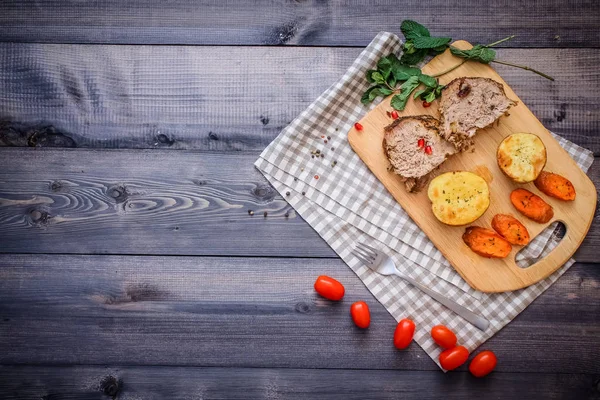 Kesme tahtası üzerinde pişmiş et iki dilim, iki dilim patates ve havuç dilimleri, keten, kareli bir peçete pişmiş bir kapta çok renkli bezelye biber ve grafit tezgahı çerçevede bir tabakta kesilmiş bir nar vardır