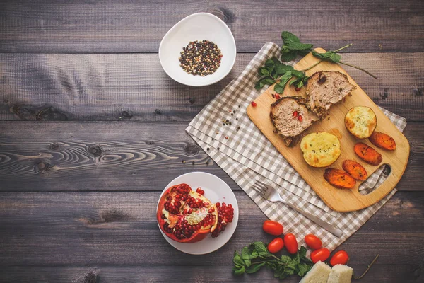 Kesme tahtası üzerinde pişmiş et iki dilim, iki dilim patates ve havuç dilimleri, keten, kareli bir peçete pişmiş bir kapta çok renkli bezelye biber ve grafit masa üstü karşı bir tabakta kesilmiş bir nar vardır