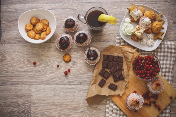 On a light wooden tabletop on a linen napkin napkin, there is a cutting board with two muffins, a broken chocolate bar and bright red berries in a small tree, next to a bowl with cookies. On a light wooden tabletop, four chocolate-covered cupcakes de