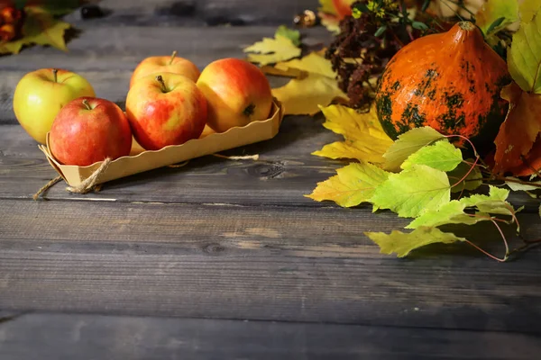 There is an orange pumpkin on a gray wooden tabletop on autumn leaves, three red apples in a wooden box.