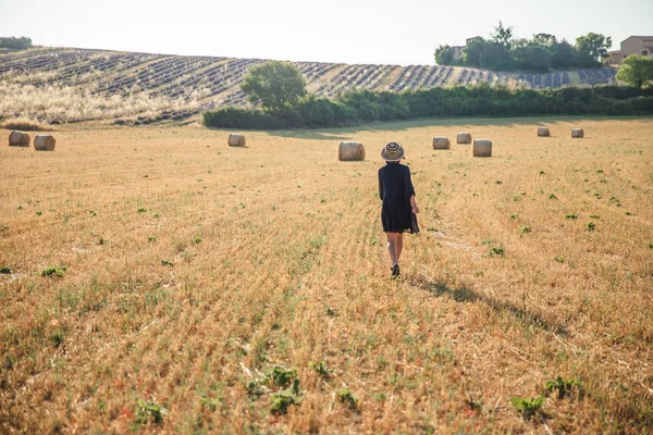 Vue arrière de fille en chapeau marchant sur le terrain agricole avec balles de foin, provence, france — Photo de stock