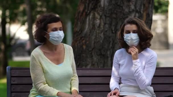 Mamma och dotter i medicinska masker pratar på en bänk. — Stockvideo