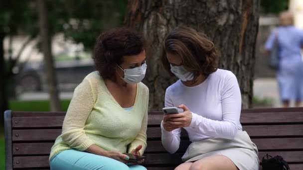 Mamá y su hija en máscaras médicas están hablando en un banco . — Vídeo de stock