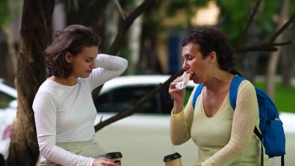 Η μαμά και η κόρη κάθονται σε ένα παγκάκι και πίνουν καφέ. — Αρχείο Βίντεο