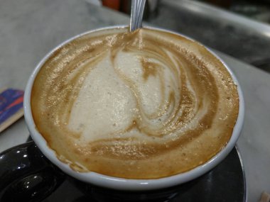 Bir fincan İtalyan kapuçino. Kremalı sıcak köpük sütünden beyaz koyu kahve kahverengi arasında değişen çizgiler vardır. Köpük karışık çay kaşığı fincan kenarında dinleniyor.