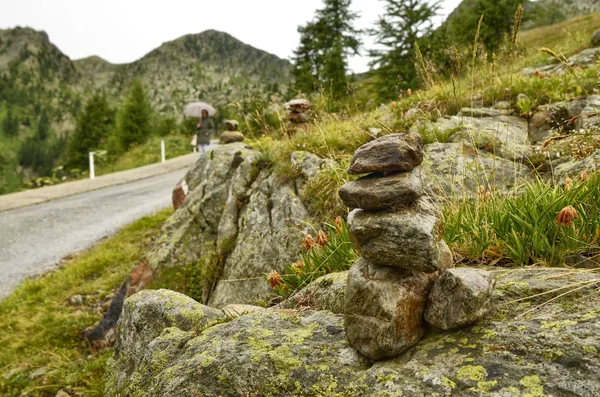 Stones piled up as a Zen memorial along a mountain road, in the background a blurred road and an indistinguishable woman.